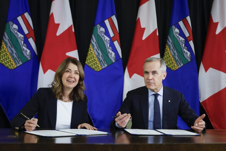 A shot of Danielle Smith and Mark Carney at a signing ceremony, with documents in front of them and pens in hand. They are both smiling toward the camera and there is a row of Alberta and Canadian flags behind them.