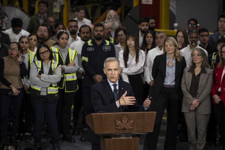 Mark Carney stands at a lectern on the floor of an auto parts plant, with workers and others gathered behind him.