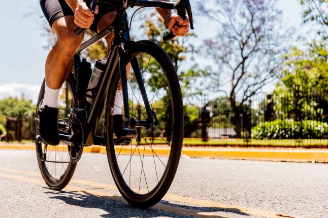 A detailed close-up capturing the dynamic moment of cycling, focusing on the cyclist's foot positioned on a bicycle pedal. The image highlights the technical cycling gear, with a crisp white sock and professional cycling shoe, set against a bright, sunlit outdoor environment.