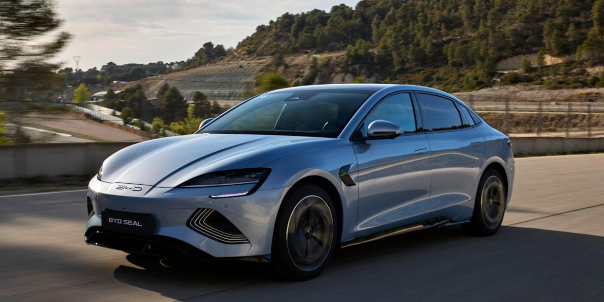 A futuristic, silver sedan drives down a road in front of green trees.
