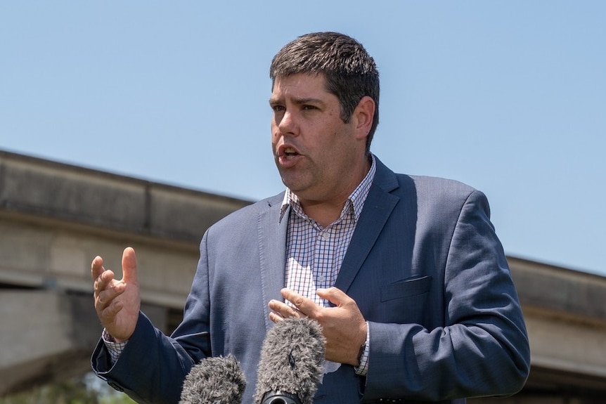 A plump 30-something man in a grey jacket gestures in front of microphones