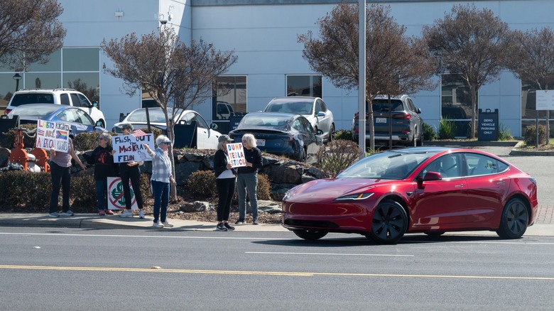 Small group of protesters holding signs outside a Tesla dealership as a red Tesla sedan drives past on the street