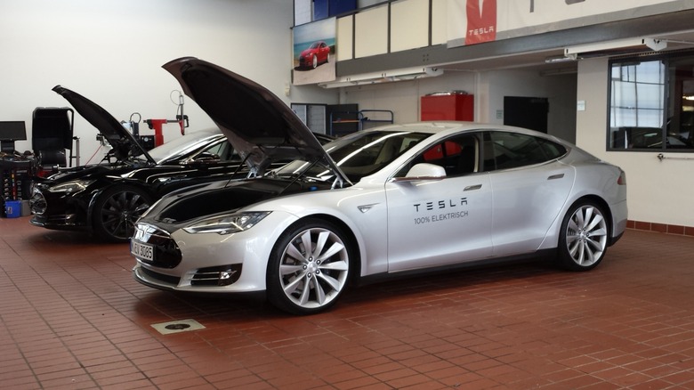 Silver Tesla sedan with its hood open inside a service garage, parked next to another Tesla vehicle