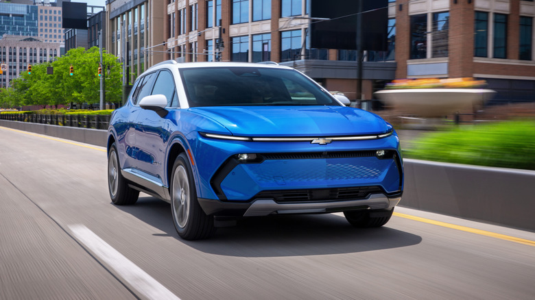 Front 7/8 view of a 2025 Chevrolet Equinox EV LT in Riptide Blue Metallic with a Summit White roof driving in a downtown area.