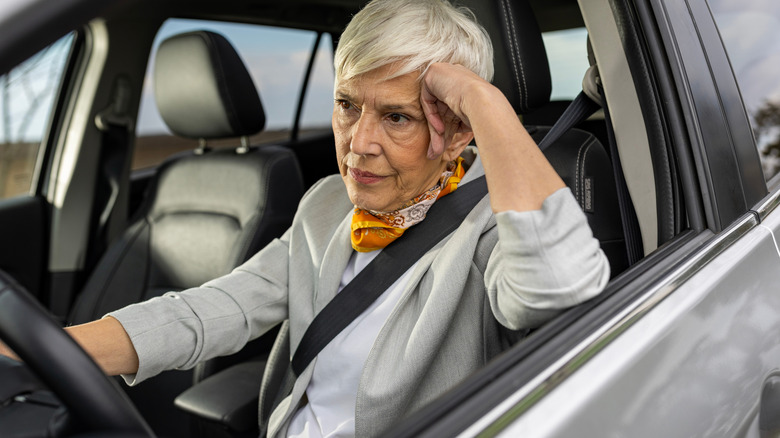 Older woman sitting in the driver's seat of a car with her seatbelt on, leaning her head on her hand