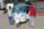 A white and blue electric auto-rickshaw stands parked, with two technicians on either side on a sunny day.
