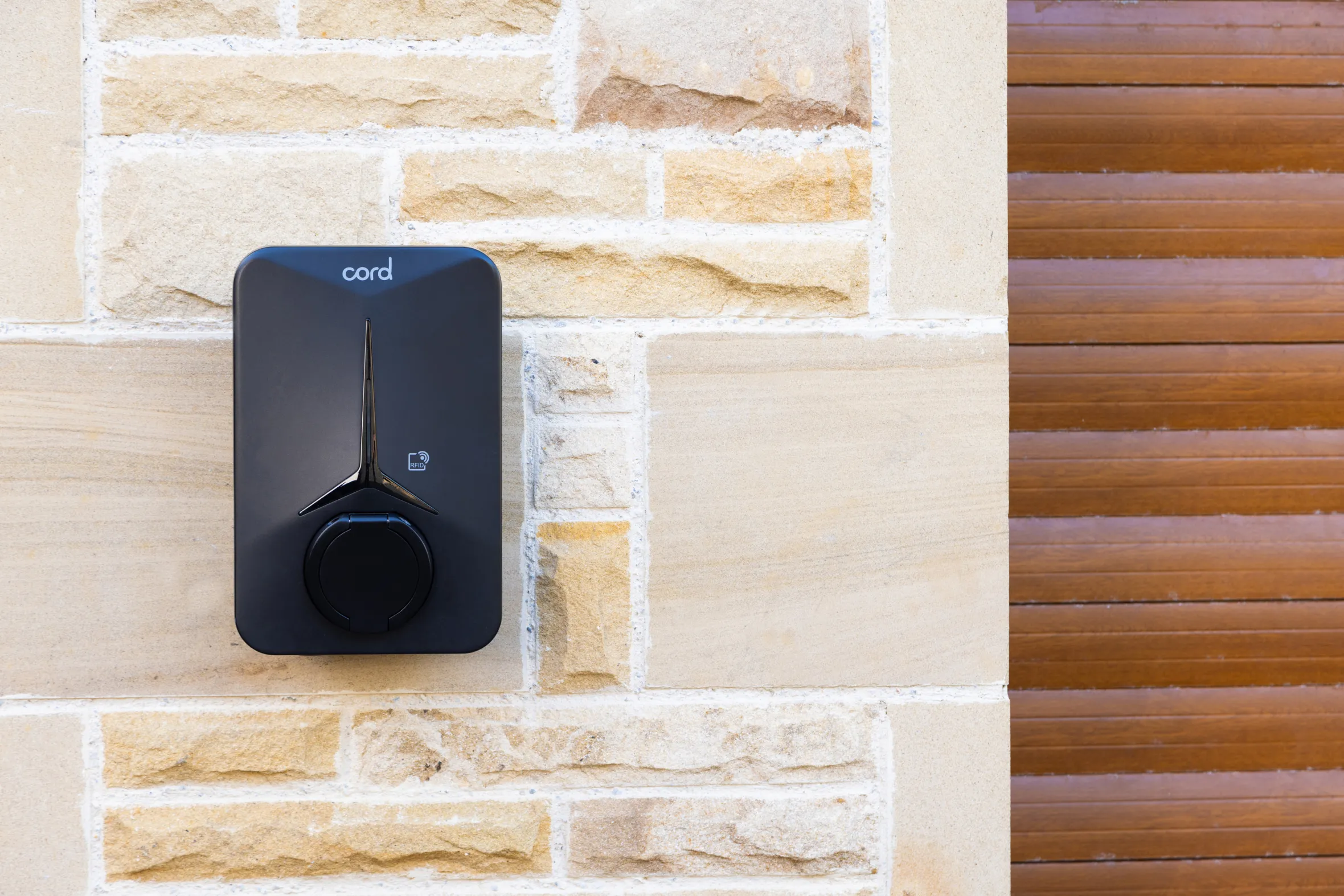 A black Cord EV charging station mounted on a light-colored brick wall next to a brown wooden garage door.