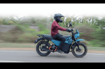 A person in a helmet and maroon shirt riding a teal electric motorcycle on a road with a blurred green background.
