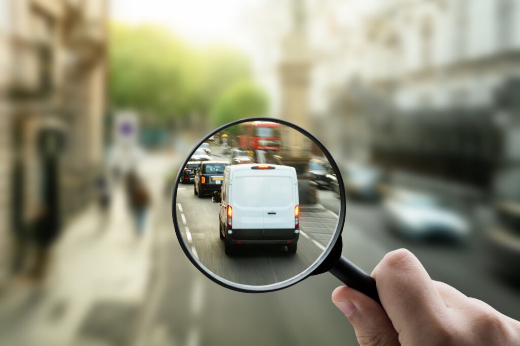 Urban street with a white delivery van driving through traffic, with a magnifying glass focusing on logistics activity.