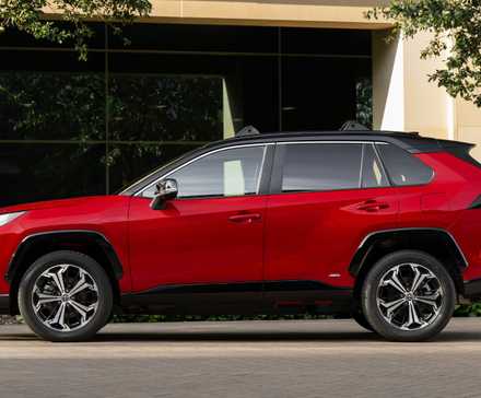 Static side profile shot of a red 2025 Toyota RAV4 PHEV XSE parked outside of a modern building.