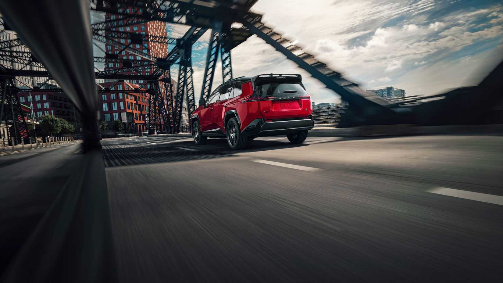 Dynamic rear 3/4 shot of a red 2026 Toyota RAV4 PHEV GR Sport driving over a bridge in a city.