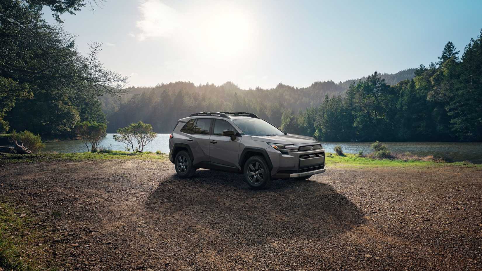Static side profile shot of a gray 2026 Toyota RAV4 Woodland parked on gravel next to a lake with trees in the background.