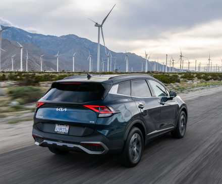 Dynamic rear 3/4 shot of a blue 2025 Kia Sportage Hybrid driving on a country road with mountains and wind turbines in the background.