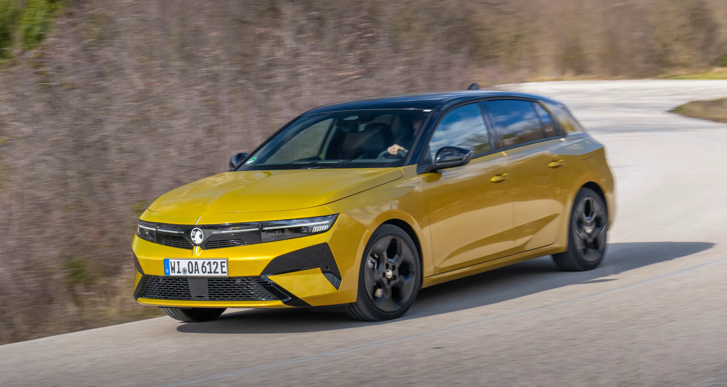 A 2026 Vauxhall Astra Hatch, yellow with a black roof, drives on a road with trees in the background.