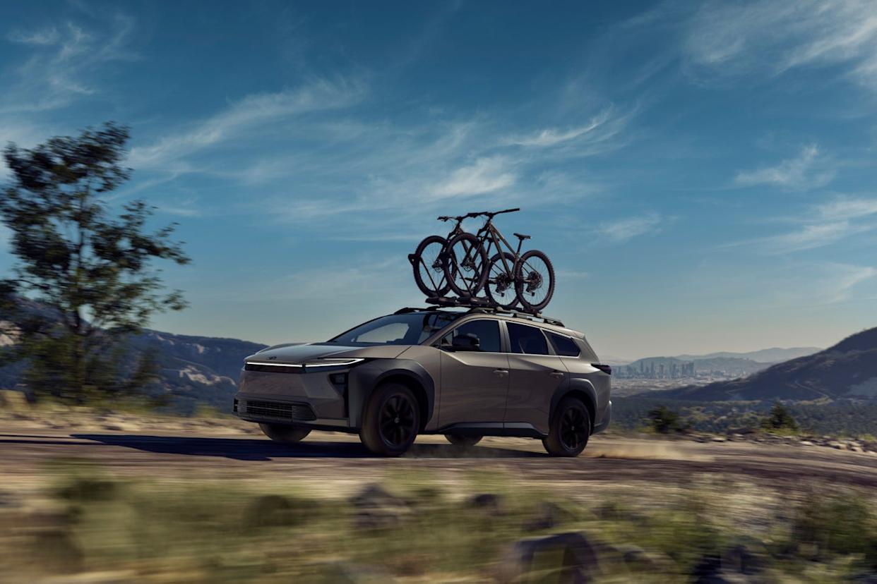 a gray suv with two bicycles mounted on the roof rack driving on a dirt road, with mountains and a cityscape in the background.