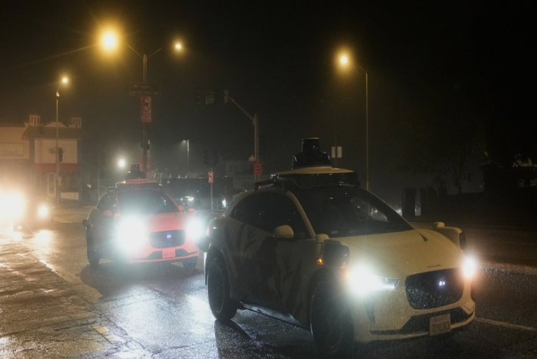 A white car with a rooftop sensor drives ahead of a police car with bright headlights on a wet street at night under streetlights.