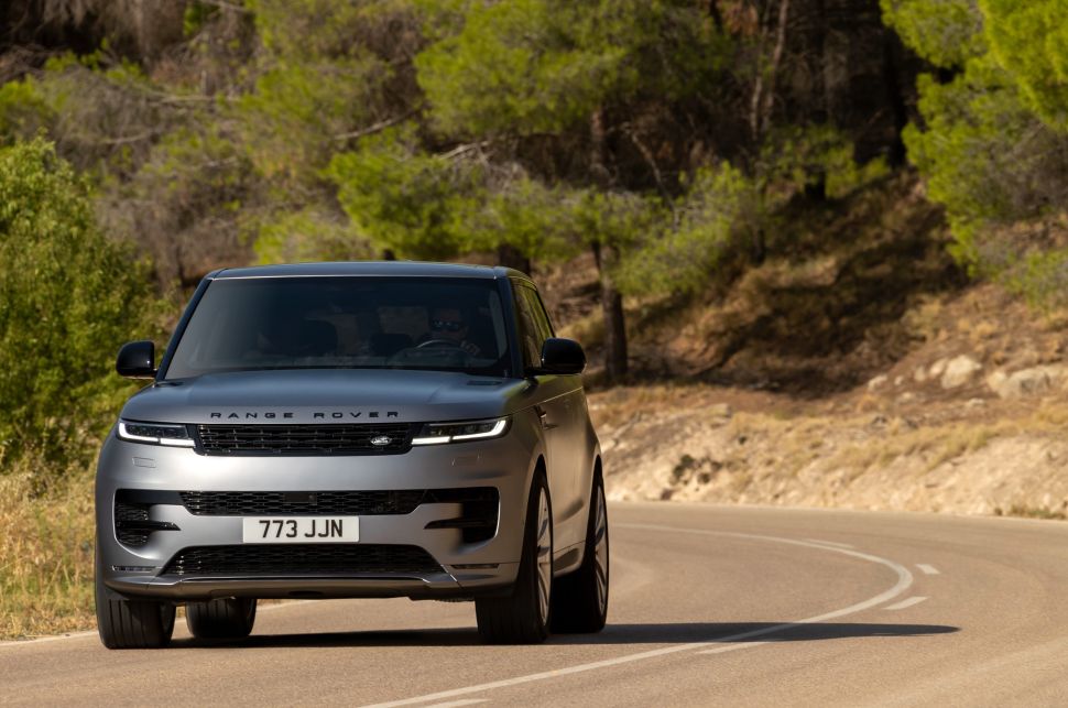 A dynamic shot of a silver Range Rover driving on a winding road, with trees and hills in the background, capturing the vehicle in motion.