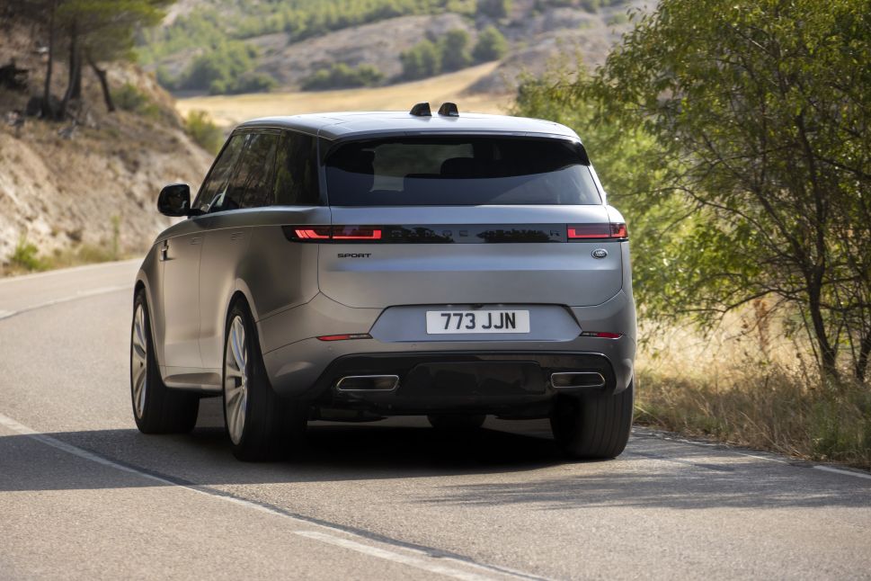 A rear view of a silver Range Rover on the road, with its license plate visible and the landscape of greenery and hills in the background.
