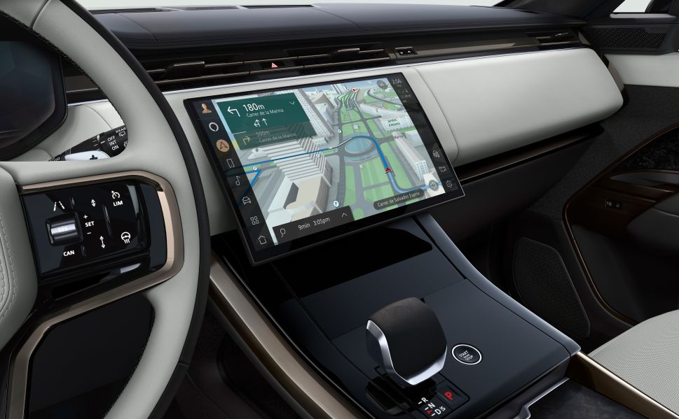 An interior view of a car showing a large touchscreen with navigation directions on display, set in the dashboard with the steering wheel and gear lever visible in the foreground.