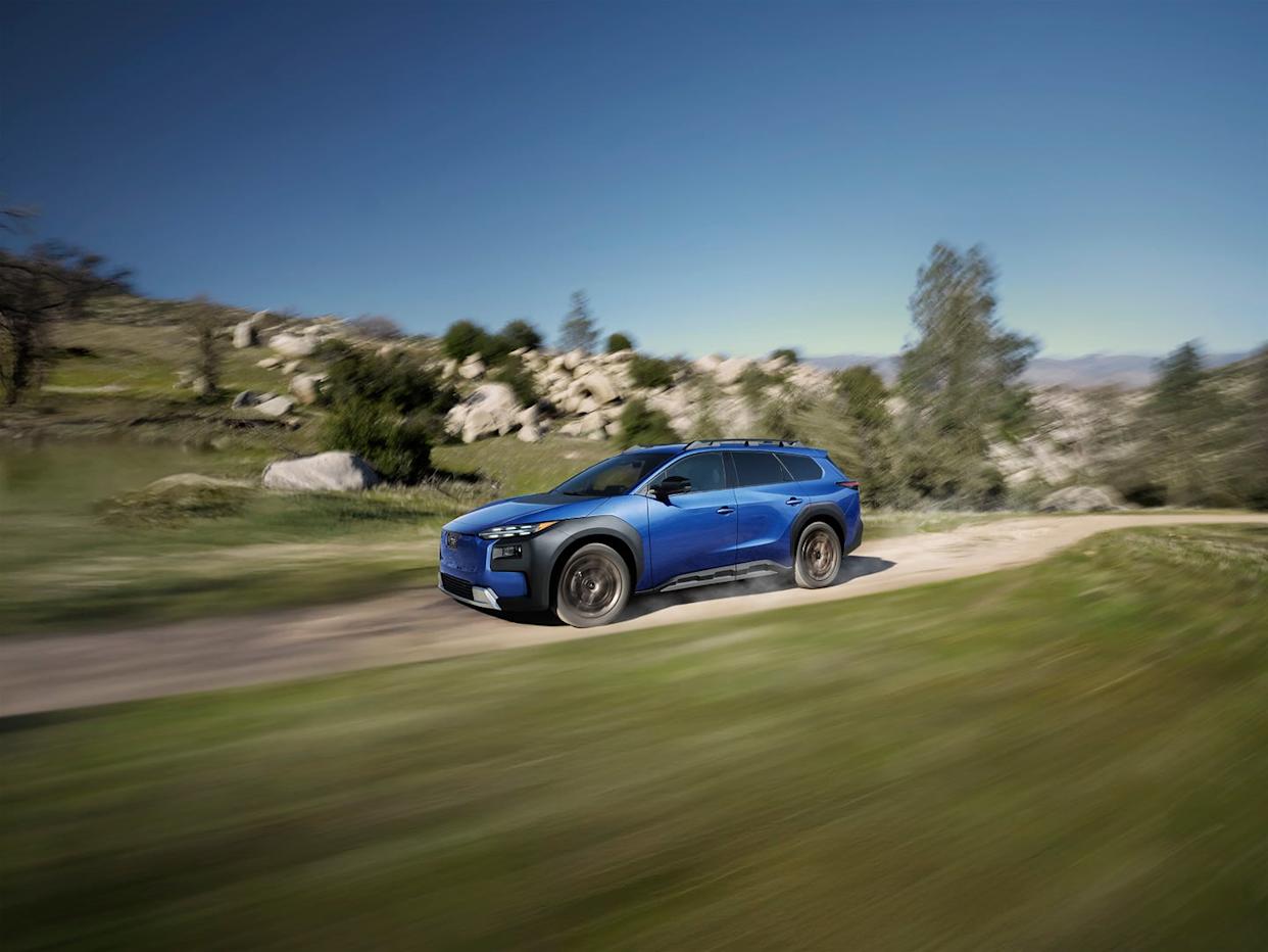 a blue suv in motion on a dirt road surrounded by greenery, rocks, and trees under a clear blue sky.