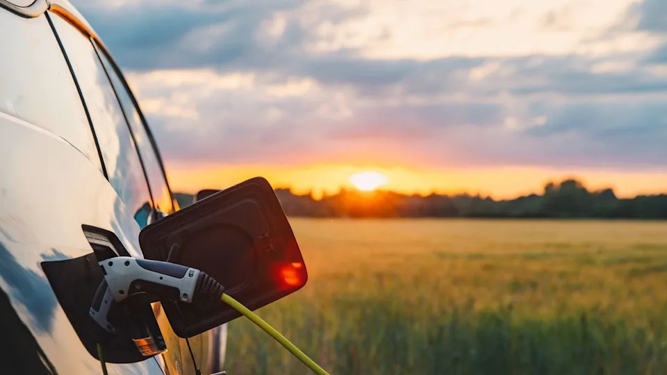  Car charging port open with a charging cable attached. the car is in an open field and the sun is setting on the horizon. 