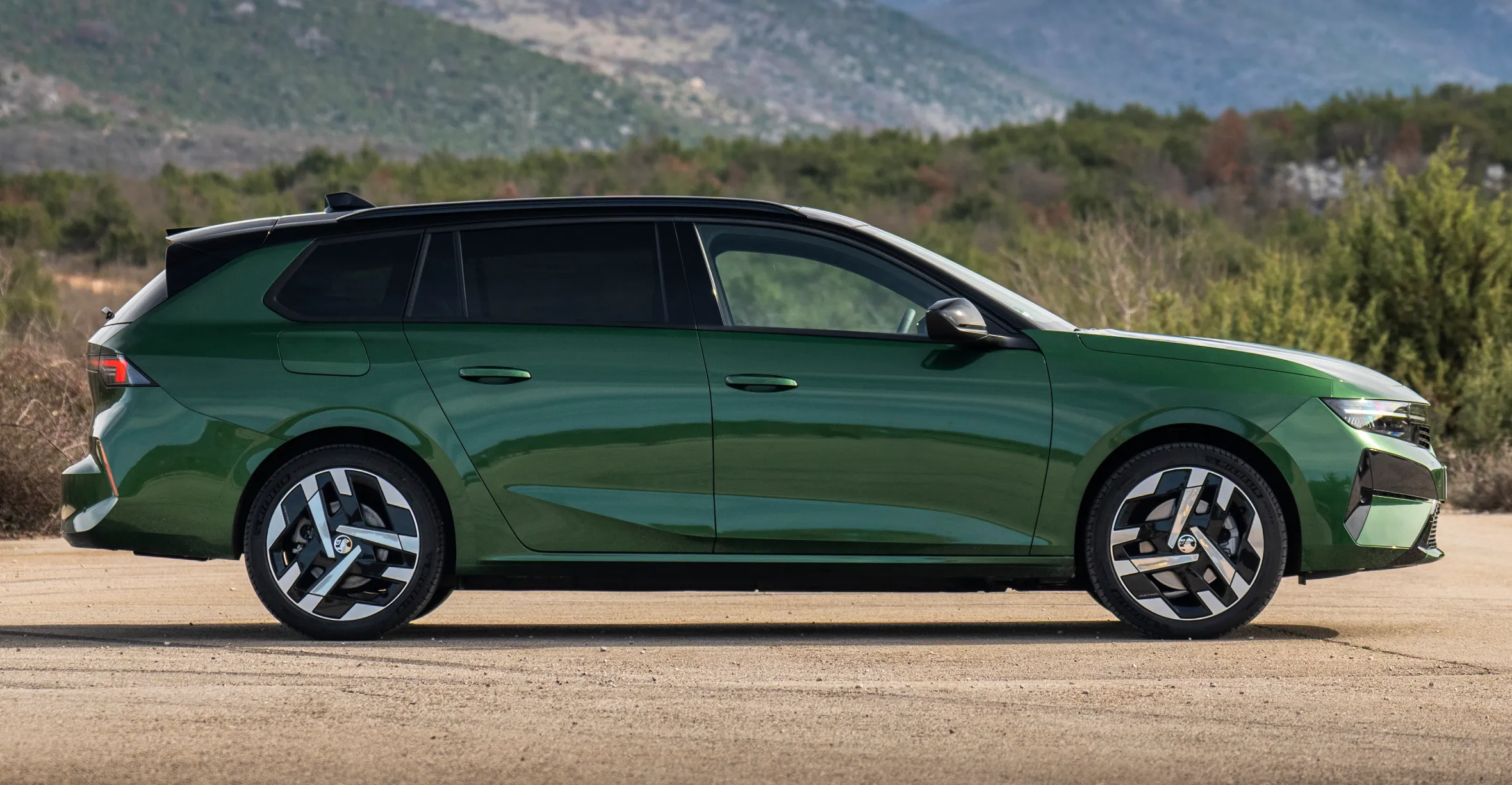 A green 2026 Vauxhall Astra Sport Tourer parked on a paved road with hills and mountains in the background.