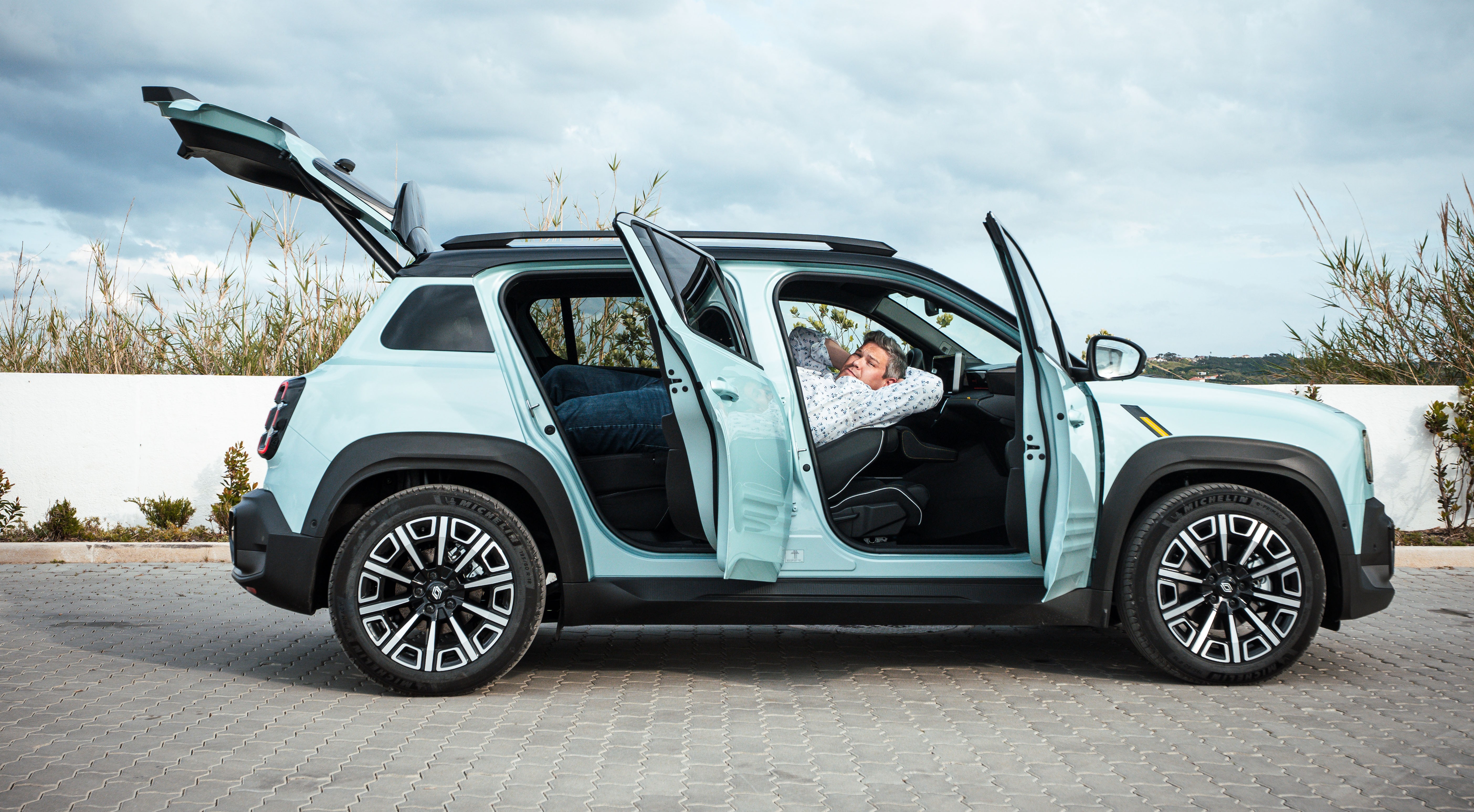 Will Dron relaxing in the front seat of a light blue Renault 4 E-Tech with all doors and the trunk open.
