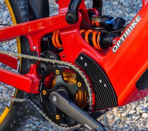 Detailed close-up of the Optibike G2 Altus electric mountain bike frame showing the red carbon fiber chassis, FOX rear shock with coil spring, integrated mid-drive motor, and high-performance chainring and crankset.