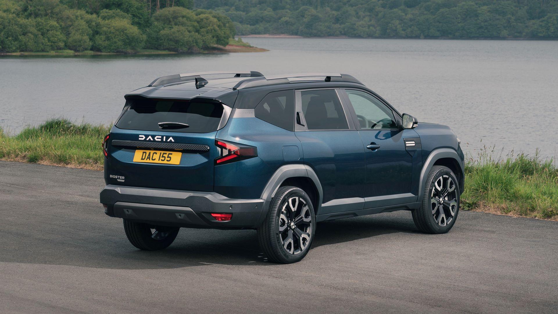 A blue Dacia Bigster parked next to a lake in the countryside, viewed from the rear.