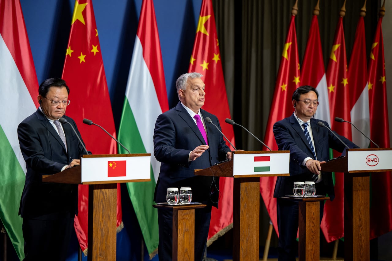 Three men stand at podiums. Each podium has a symbol: the flag of China, the flag of Hungary, and the logo of EV car company BYD.