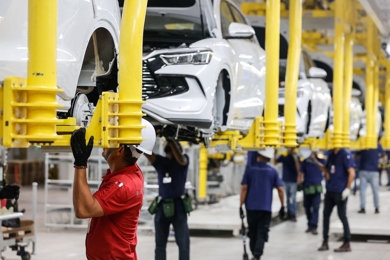 Cars under construction are suspended from machinery in a factory while workers stand by.
