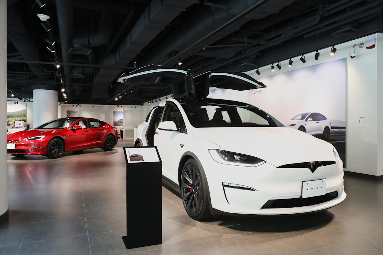 A white Tesla Model X is parked in front of a red Tesla Model S on a showroom floor.