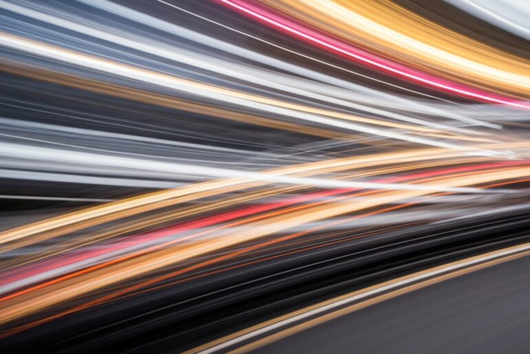 An abstract, colorful image created with a slow shutter speed and panning motion, depicting a vehicle in motion as a series of vibrant, blurred streaks, conveying a sense of speed and energy without any identifiable details.