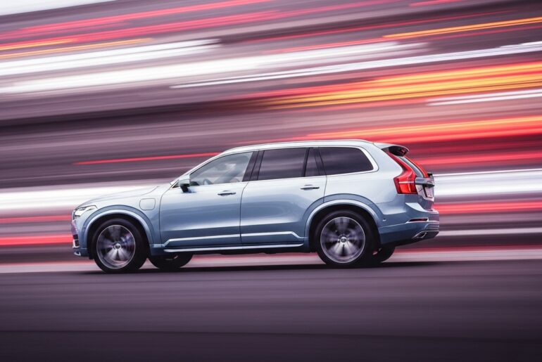 An abstract, sweeping image of a Volvo XC90 plug-in hybrid SUV, its form blurred into vibrant streaks of color, conveying a sense of speed and modern travel.