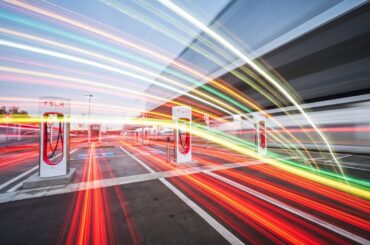 An abstract, colorful image created with a slow shutter speed and panning motion, depicting a Tesla Supercharger station as sweeping, blurred lines of vibrant hues, conveying the dynamic energy and modern technology of electric vehicle charging.