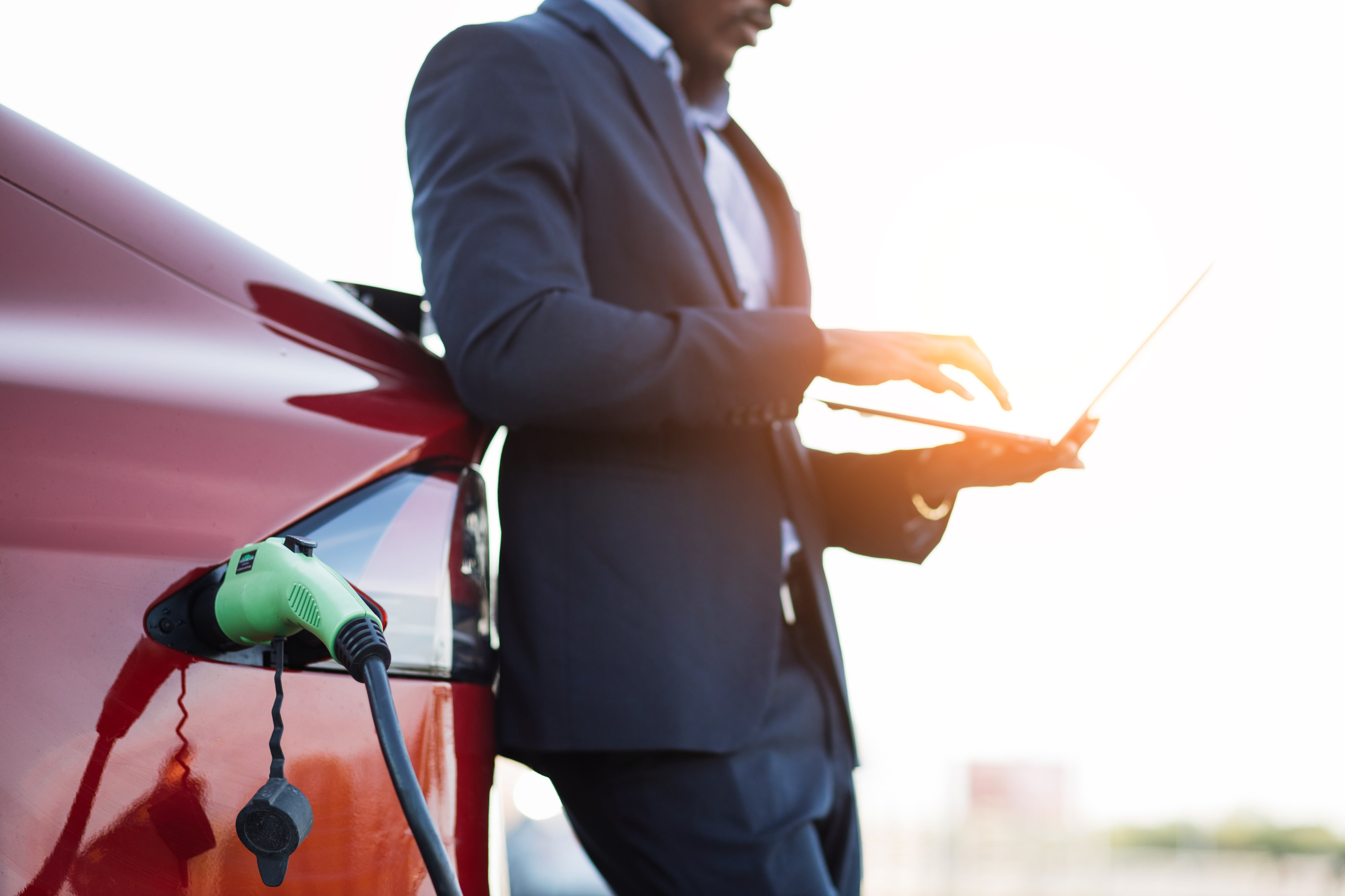 A man uses a laptop while leaning against his EV, which is charging.
