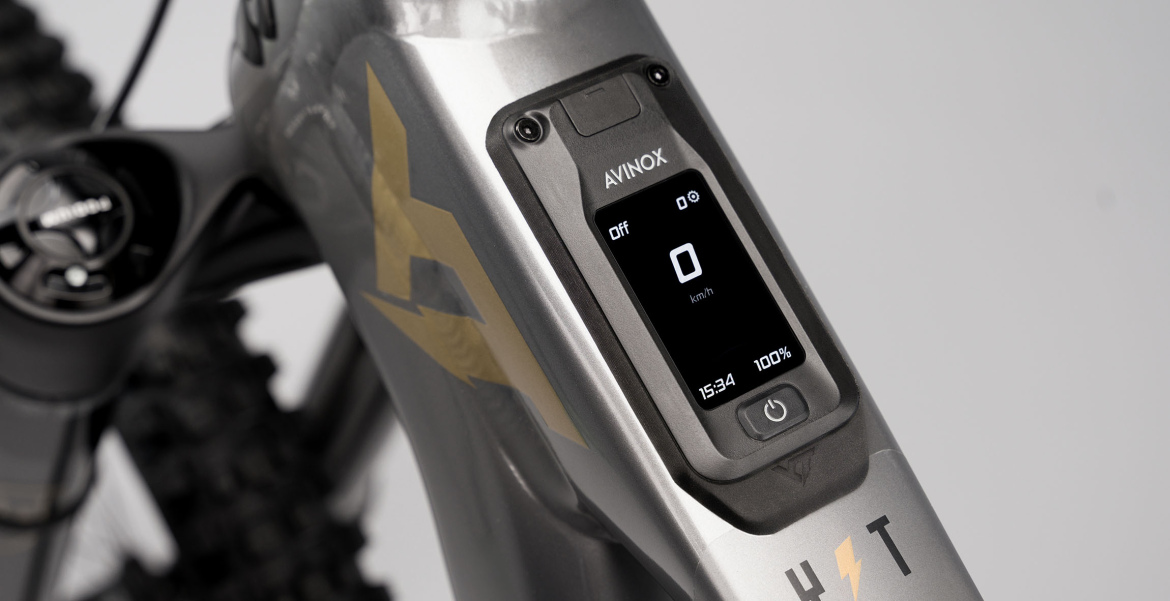Close-up of a sleek electric bike display, showing speed as "0 km/h," battery status at "100%," and time as "15:34." The bike frame features a metallic gray finish with decorative gold accents.