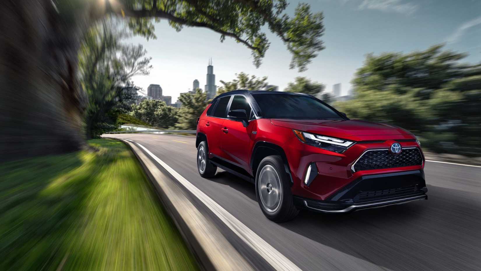 Dynamic front 3/4 shot of a red 2024 Toyota RAV4 Prime XSE driving on a road lined with trees with a cityscape in the background.