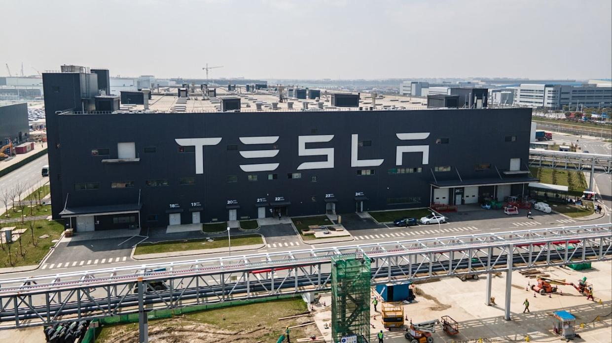 An aerial, eye-level shot of the dark gray Tesla Gigafactory building in Shanghai, China, with a large white 'TESLA' logo stretching across its side. Below the logo, a series of loading docks are numbered from 18 to 26. In the foreground, a gray metal pedestrian bridge or conveyor system extends across the frame, with construction activity and workers visible on the ground beneath it. The background shows an extensive industrial complex under a bright, slightly hazy sky.