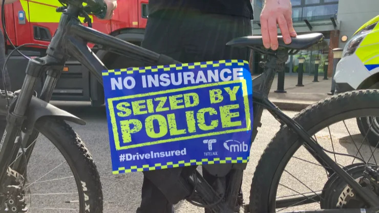The close-up image shows a banner or poster on an e-bike which says: "No insurance. Seized by police" in green letter on a blue background. The e-bike is black and is being held upright by a police officer.