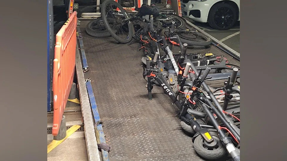 A large collection of e-bikes and e-scooters on the back of a lorry, on their side. A white car is to the left and an orange barrier to the right. It looks like the items are on a ramp. 