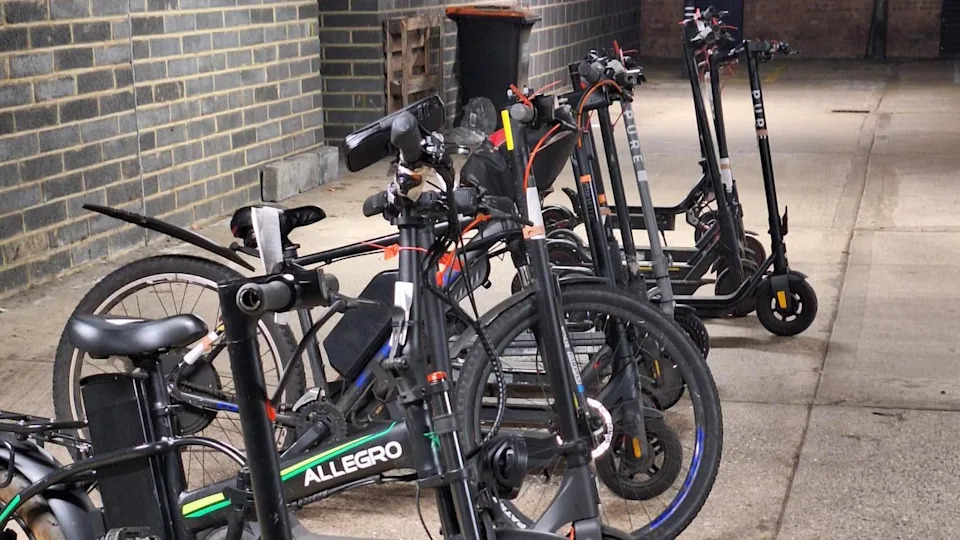 A row of black e-scooter and e-bikes in a room, that has grey brick walls and a dark floor. They are all standing up with a wheelie bin in the distance, against the wall. 