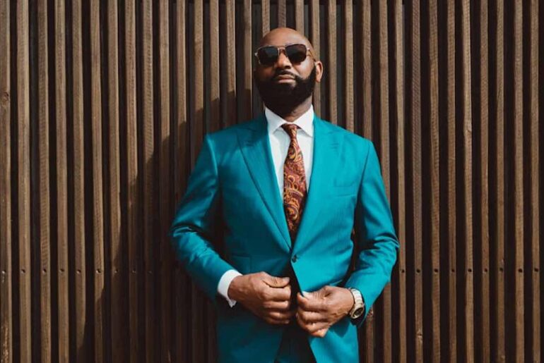 A handsome mature bald bearded black man in a sunglasses and a fashionable blue or teal costume with a tie is standing in front of a wall made of striped wooden timbers and fastening a suit button