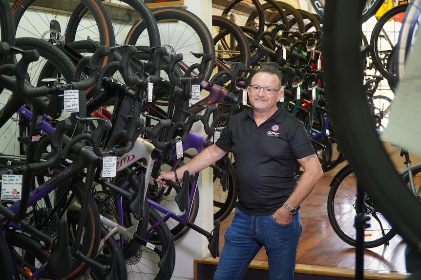 A man wearing a black shirt and blue jeans stands in a bike shop posing for a photo.