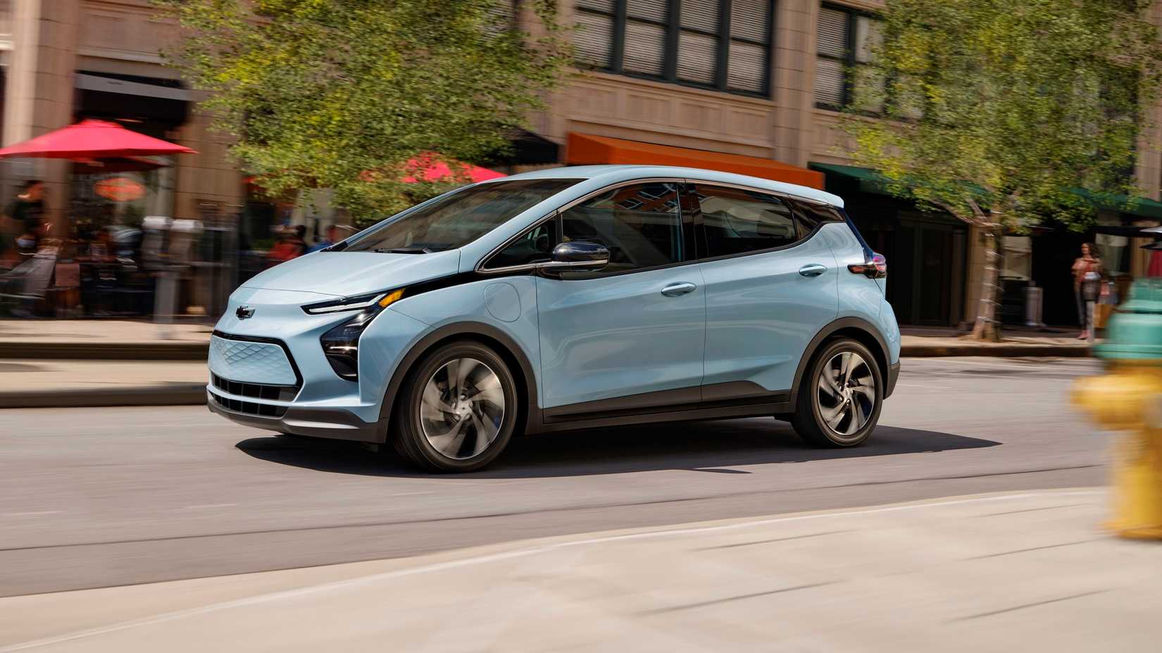 A dynamic front-quarter tracking shot of a blue Chevrolet Bolt EV driving down a city.