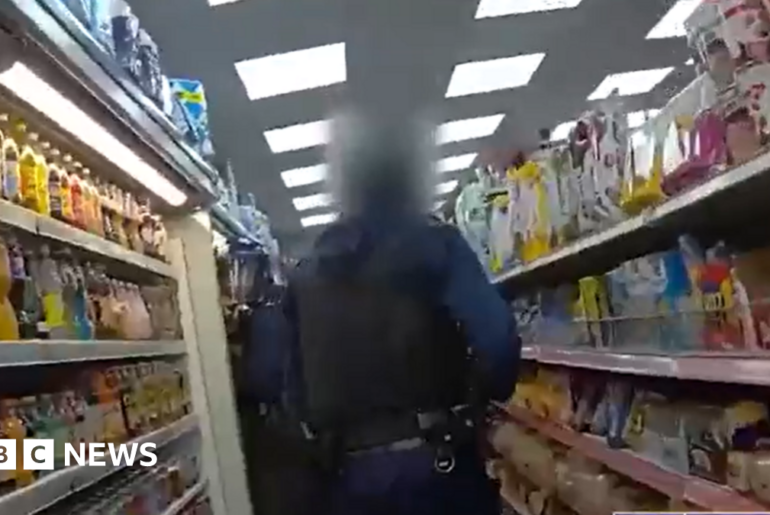 Still image taken from bodycam footage. Two police officers in dark clothing are in view from behind as they walk down the food aisle of a shop. There are food products on the shelves on either side of the officers.