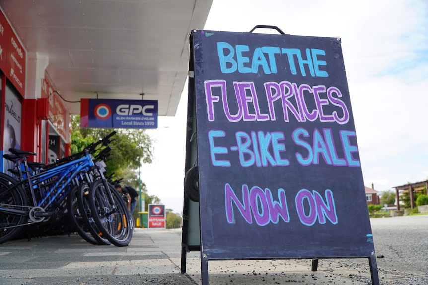 A sign on the ground outside a bike shop reading "beat the fuel prices, e-bike sale now on".