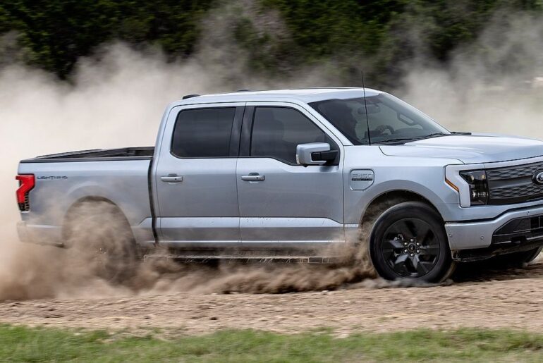 Silver Ford F-150 Lightning Lariat electric pickup truck drifting sideways on dirt rally course with large dust cloud