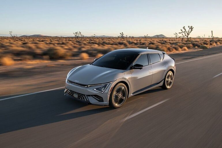 Silver Kia EV6 electric crossover cruising desert highway at sunset with Joshua trees in background