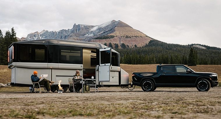 An image of Lightship's EV RV AE.1 being towed by a black pickup truck, with a couple camping outside the travel trailer.